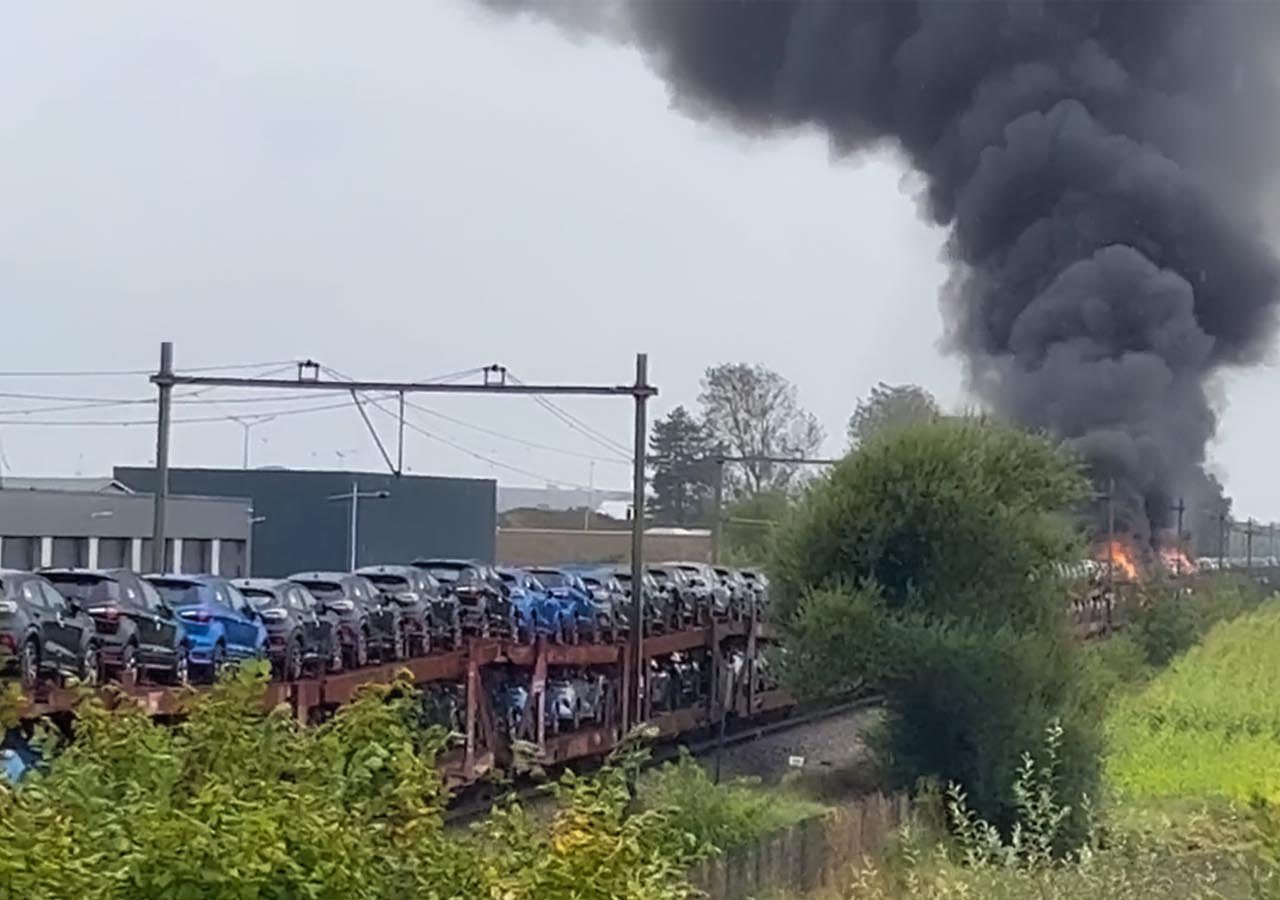 In de verte is een grote brand zichtbaar met dikke zwarte rook die opstijgt, grenzend aan een goederentrein beladen met voertuigen op meerdere autotransportwagens.