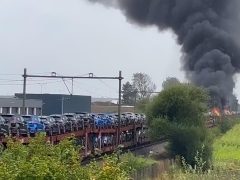 In de verte is een grote brand zichtbaar met dikke zwarte rook die opstijgt, grenzend aan een goederentrein beladen met voertuigen op meerdere autotransportwagens.