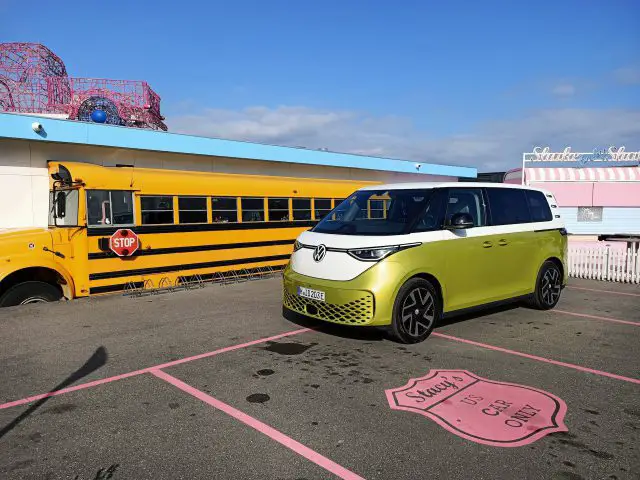 Een gele Volkswagen ID. Buzz staat op een zonnige dag geparkeerd op een parkeerplaats met een gele schoolbus en een gebouw op de achtergrond, wat de essentie van een Volkswagen ID Buzz-test vastlegt.