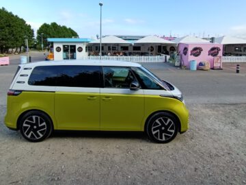 Een groen-witte Volkswagen ID Buzz-testminibus staat geparkeerd voor een roze en wit gebouw met zitplaatsen buiten en bomen op de achtergrond.