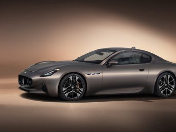 Een slanke, grijze tweedeurs Maserati GranTurismo-sportwagen met een soepel, gewelfd ontwerp wordt tentoongesteld onder een spotlight in een neutraal getinte studioomgeving.