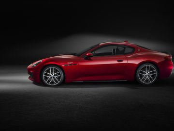 Een zijaanzicht van een rode Maserati GranTurismo in een slecht verlichte omgeving met een spotlight die het voertuig benadrukt. De auto beschikt over strakke aerodynamische lijnen en zilveren wielen, die elegantie en prestaties uitstralen.