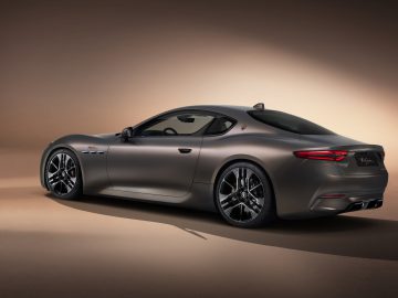 Een slanke, grijze Maserati GranTurismo-sportwagen wordt gepresenteerd in een slecht verlichte studio met spotlight erop, wat het aerodynamische ontwerp en de moderne kenmerken benadrukt.