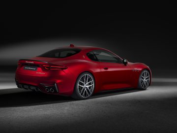 Linksachter is een rode Maserati GranTurismo-sportwagen te zien, met strakke designlijnen, viervoudige uitlaatpijpen en lichtmetalen velgen, verlicht tegen een donkere achtergrond.