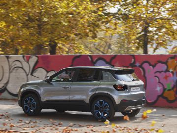 Een zilveren Jeep Avenger rijdt langs een levendige muurschildering met graffitikunst en herfstbladeren op de achtergrond.