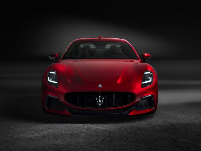 Een rode Maserati GranTurismo wordt verlicht tegen een donkere achtergrond, waardoor het vooraanzicht met de koplampen aan en het Maserati-logo op de grille goed zichtbaar is.