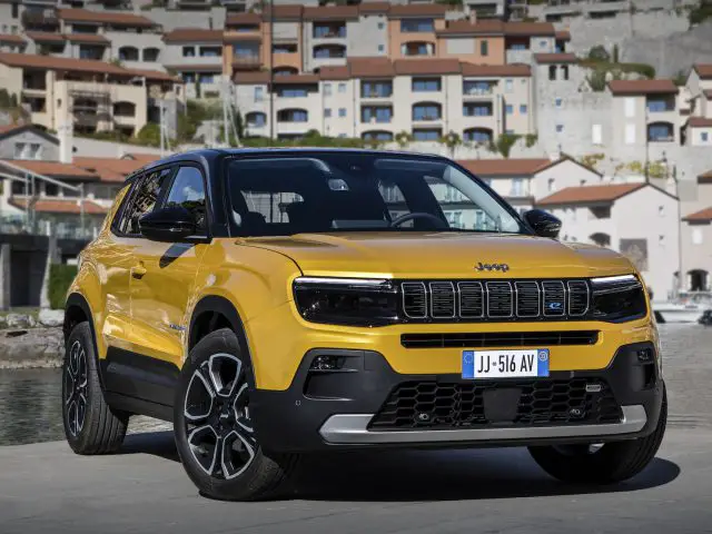 Een gele Jeep Avenger SUV staat geparkeerd vlakbij een waterkant met op de achtergrond gebouwen met meerdere verdiepingen en boten.