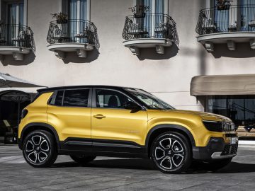 Een gele Jeep Avenger staat geparkeerd voor een beige gebouw met balkons en grote ramen.