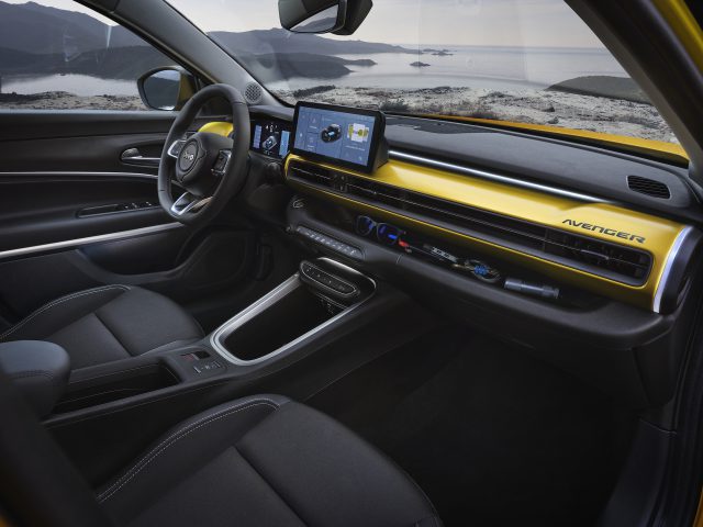 Interieur van een moderne Jeep Avenger met een zwart en geel dashboard met het woord 'Avenger', een aanraakscherm en zwarte stoelen. Op de achtergrond is door de ramen een schilderachtig uitzicht op een meer en de heuvels zichtbaar.