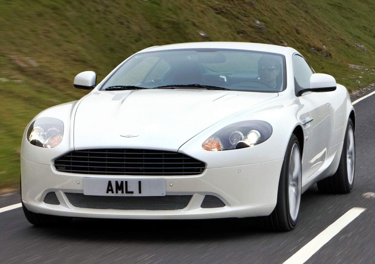 Een witte Aston Martin DB9 met kenteken "AML 1" rijdt op een weg met met gras begroeide heuvels op de achtergrond.
