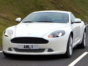 Een witte Aston Martin DB9 met kenteken "AML 1" rijdt op een weg met met gras begroeide heuvels op de achtergrond.