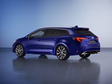 Een blauwe Toyota Corolla Touring Sports Hybrid Wagon, gezien vanaf de linkerachterkant, tegen een gradiëntblauwe achtergrond.
