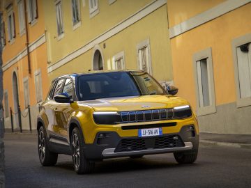 Een gele Jeep Avenger SUV staat geparkeerd in een smalle, lege straat vol levendige gele en oranje gebouwen.