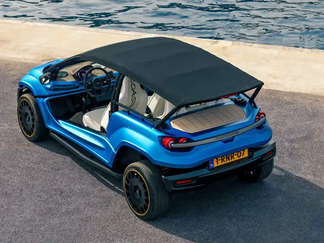 Een blauwe compacte Coastrunner EV met een zwart softtopdak staat aan het water geparkeerd op een betonnen ondergrond. Het elektrische voertuig heeft een open ontwerp met twee stoelen en een zichtbaar stuur.
