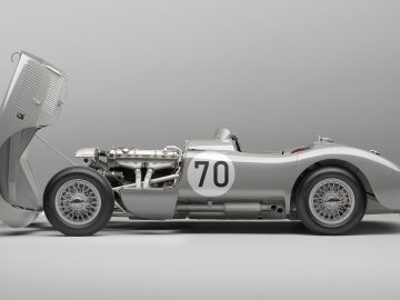 Een vintage zilveren Jaguar C-Type raceauto met het nummer 70 op de zijkant, in profielaanzicht, met de voorkap open waardoor de motor zichtbaar is.