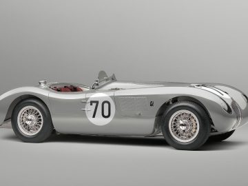 Zijaanzicht van een klassieke zilveren Jaguar C-Type raceauto met het nummer 70 op de deur tegen een effen grijze achtergrond.