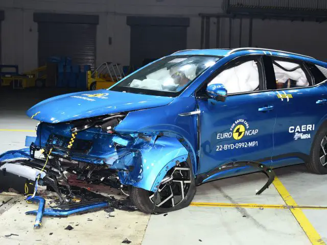 Een blauwe BYD Atto 3 SUV met ernstige frontale schade die een crashtest ondergaat in een binnenfaciliteit. De Euro NCAP- en CAERI-logo's zijn zichtbaar op het voertuig. Het puin ligt verspreid over de auto.