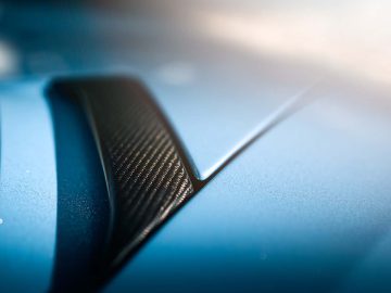 Close-up van de motorkap van een Aventador met een blauw metallic afwerking en een zwarte koolstofvezel ventilatieopening onder fel licht.