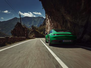 Een groene Porsche 911 Carrera T rijdt op een zonnige dag over een kronkelende bergweg met rotswanden en een bergachtig landschap op de achtergrond.