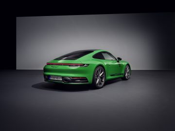 Een strakke, groene Porsche 911 Carrera T staat geparkeerd in een goed verlichte, minimalistische setting. De auto is van achteren gezien en laat het aerodynamische ontwerp en het zwarte dak goed zien.