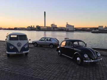 Drie klassieke auto's, waaronder een Volkswagen-bus die doet denken aan het iconische ontwerp van Ben Pon, een zilveren sedan en een zwarte Volkswagen-kever, geparkeerd aan de waterkant bij zonsondergang met industriële gebouwen op de achtergrond.