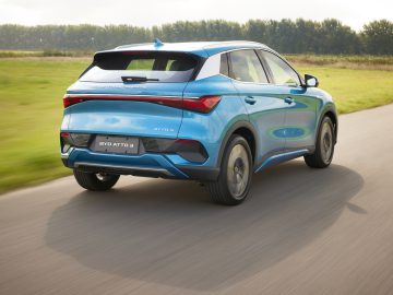 Een blauwe BYD Atto 3 elektrische SUV rijdt over een verharde landweg, omgeven door weelderige groene velden en bomen op de achtergrond.