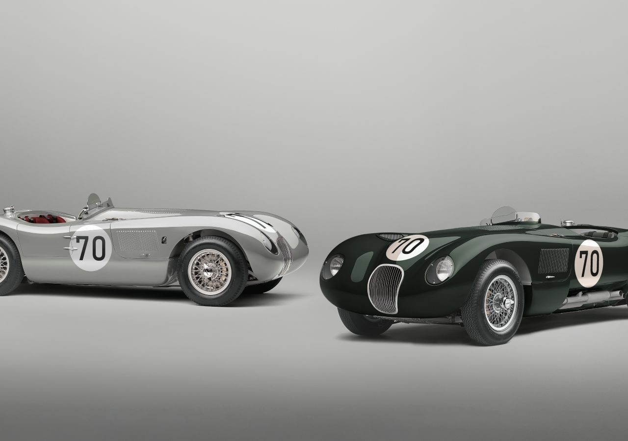 Twee vintage racewagens met nummer 70 op de zijkanten. De ene is een zilveren Jaguar C-Type en de andere is donkergroen, beide tegen een effen grijze achtergrond.