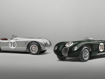Twee vintage racewagens met nummer 70 op de zijkanten. De ene is een zilveren Jaguar C-Type en de andere is donkergroen, beide tegen een effen grijze achtergrond.