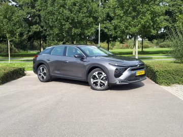 Een strakke grijze Citroën C5 X geparkeerd op een asfaltoppervlak met groen en bomen op de achtergrond, met het stijlvolle Nederlandse kenteken op de voorkant.