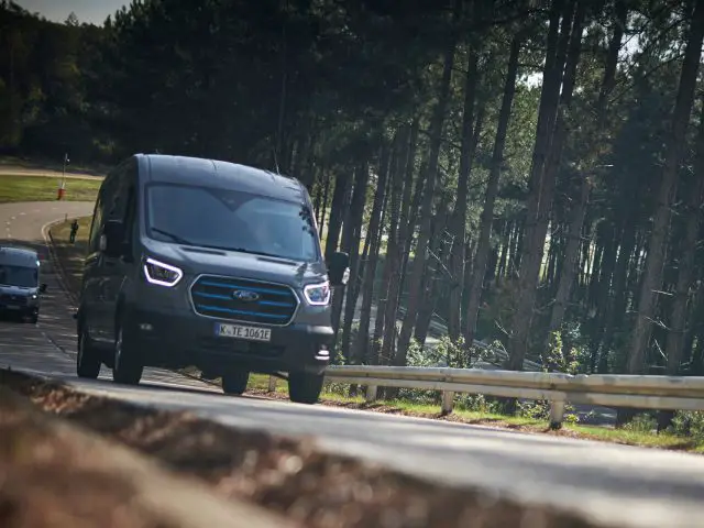 Twee voertuigen rijden op een bochtige weg door een bosrijk gebied, met de focus op een donkerblauwe Ford E-Transit op de voorgrond. Hoge bomen staan langs de kant van de weg en zonlicht filtert door het gebladerte.