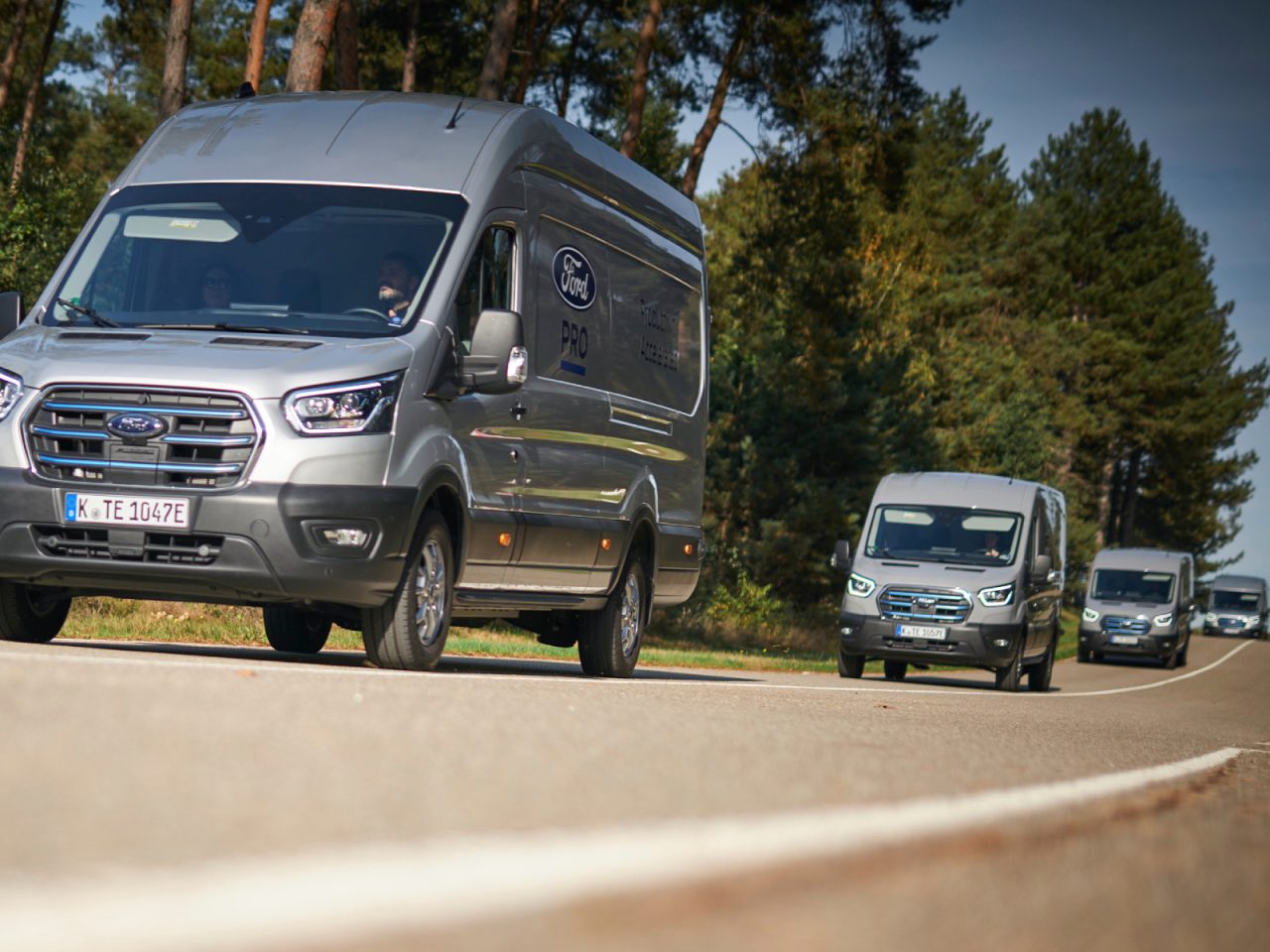 Vier zilverkleurige Ford E-Transit-busjes rijden onder een heldere hemel over een landelijke weg omzoomd met bomen.
