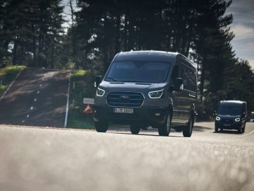 Twee zwarte Ford E-Transit-busjes rijden op een weg omgeven door hoge bomen, met één busje op de voorgrond en een ander iets daarachter. In de verte buigt de weg omhoog.