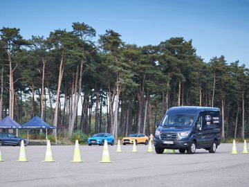 Een zwarte Ford E-Transit manoeuvreert door verkeerskegels in een open gebied met twee auto's en bomen op de achtergrond. Aan de linkerkant van de afbeelding zijn twee tenten te zien.