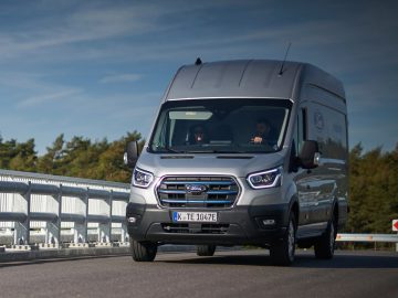 Een grijs Ford E-Transit-busje rijdt op een snelwegbrug met een bewolkte lucht boven zijn hoofd. Voorin zitten twee personen.