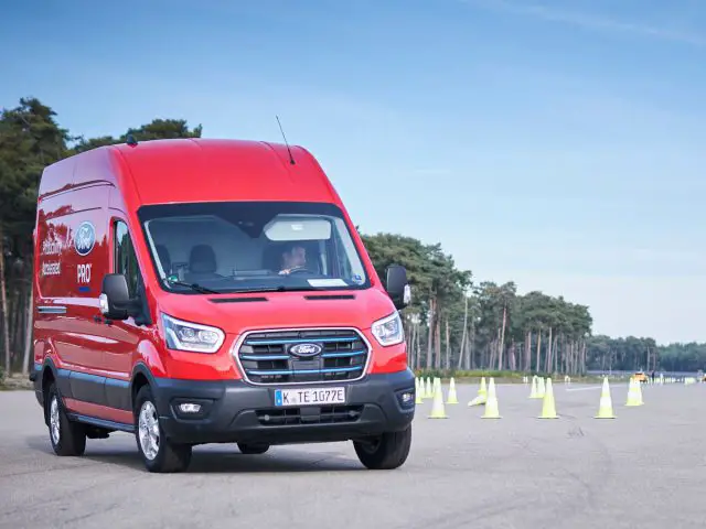 Een rood Ford E-Transit-busje rijdt op een verharde weg omringd door verkeerskegels, met bomen op de achtergrond.