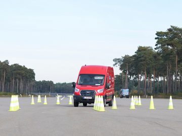 Een rood Ford E-Transit-busje navigeert door een kegelvormige hindernisbaan op een brede weg met aan weerszijden bomen.