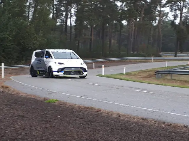 Een witte Ford E-Transit bestelwagen met diverse emblemen rijdt een bocht door over een bosrijke weg.