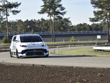 Een witte Ford E-Transit performance bestelwagen rijdt op een verharde weg met bomen en een gebogen structuur op de achtergrond.