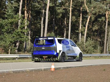 Een op maat gemaakte Ford E-Transit-bestelwagen rijdt op een weg omgeven door bos, met wit en blauw geverfde delen, en een veiligheidskegel zichtbaar op de voorgrond.