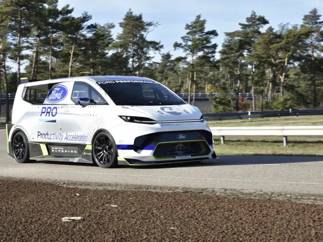 Een wit-blauwe Ford E-Transit-prestatiebestelwagen met het opschrift "Pro" en "Productivity Accelerators" rijdt op een verharde weg omgeven door bomen.