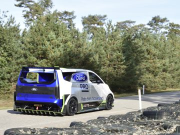 Een aangepaste Ford E-Transit-bestelwagen met racestickers rijdt op een weg omringd door pijnbomen, met stapels banden langs de weg. Het busje is voorzien van het merk Ford Pro en Product met Acceleration-logo's.