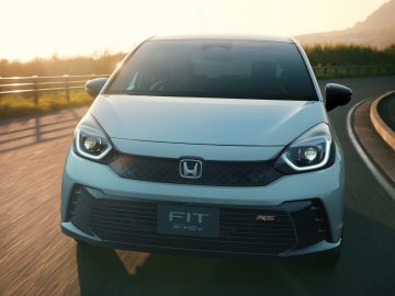 Een witte Honda Jazz RS e:HEV rijdt op een weg met een gebogen vangrail, onder een heldere hemel tijdens zonsondergang.