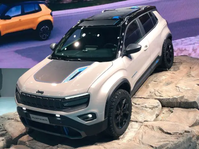 Op Autosalon Parijs 2022 wordt op een rotsachtig terrein een Jeep SUV getoond. Een groot scherm op de achtergrond toont een gele SUV die rijdt, terwijl er twee mensen vlakbij staan.