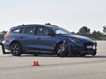 Op een open, verhard terrein met verkeerskegels staat een blauwe hatchback uitgerust met gespecialiseerde testapparatuur geparkeerd, perfect voor een rijvaardigheidstraining. Op de achtergrond zijn bomen zichtbaar.
