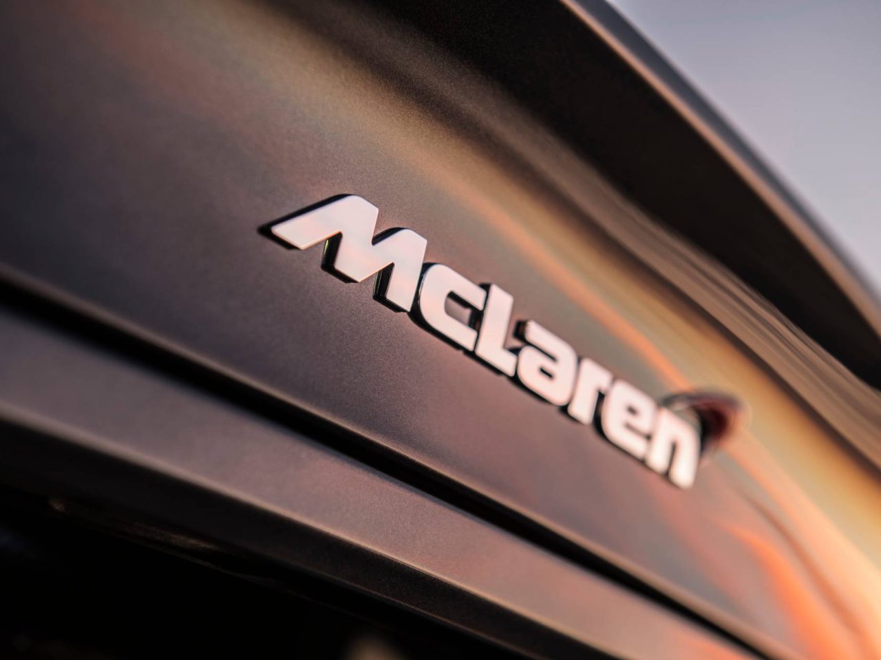 Close-up van de achterkant van een zwarte McLaren SUV, met het logo prominent weergegeven tegen een bij zonsondergang verlichte achtergrond.