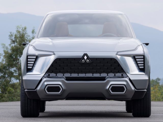 Vooraanzicht van de Mitsubishi XFC Concept, een zilveren conceptauto met opvallende geometrische koplampen en een zwarte zeshoekige grille, geparkeerd op een weg met bergen en bomen op de achtergrond.