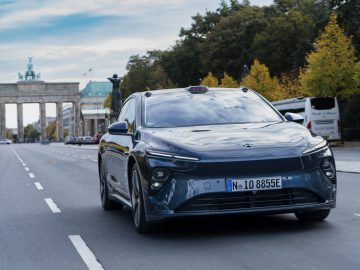 Een strakke zwarte Nio ET7 rijdt op een heldere dag over een lege weg richting de Berlijnse Brandenburger Tor, met bomen langs de straat en een bus op de achtergrond.