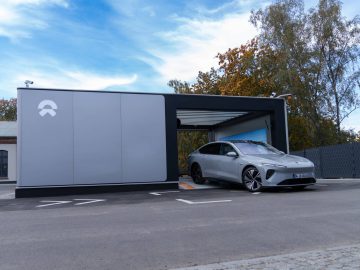 Een strakke, zilveren Nio ET7 staat geparkeerd bij een moderne, doosachtige structuur met een open deur, tegen een achtergrond van bomen en een gedeeltelijk bewolkte lucht.