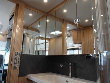 Moderne camperbadkamer met een reflecterende spiegelkast, een strak zwart aanrechtblad en een witte wastafel met een chromen kraan. Accenten op houten panelen en sfeerverlichting zijn zichtbaar.