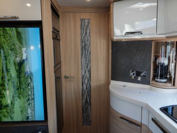 Modern camperinterieur met houten kasten, flatscreen-tv met een natuurtafereel, compacte keuken met gootsteen, fornuis en koffiezetapparaat in deze strakke camper, en een deur met een verticaal decoratief raampaneel.